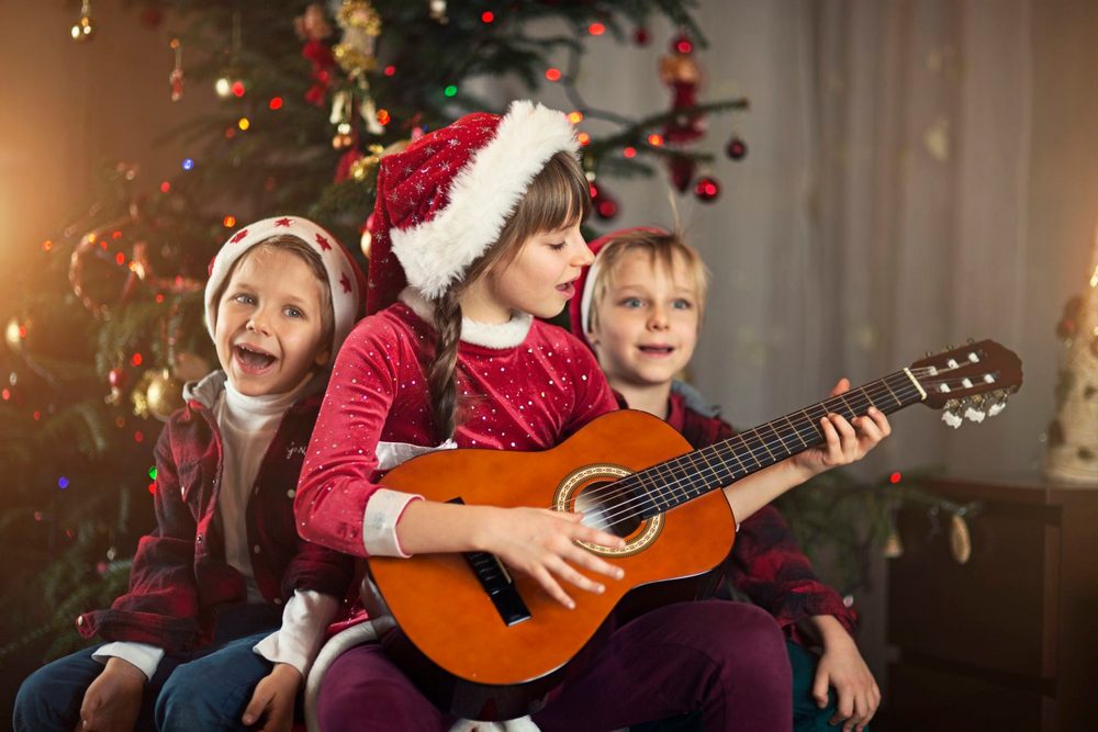 Дети с гитарой в руках в новогодних колпаках, украшенная елка