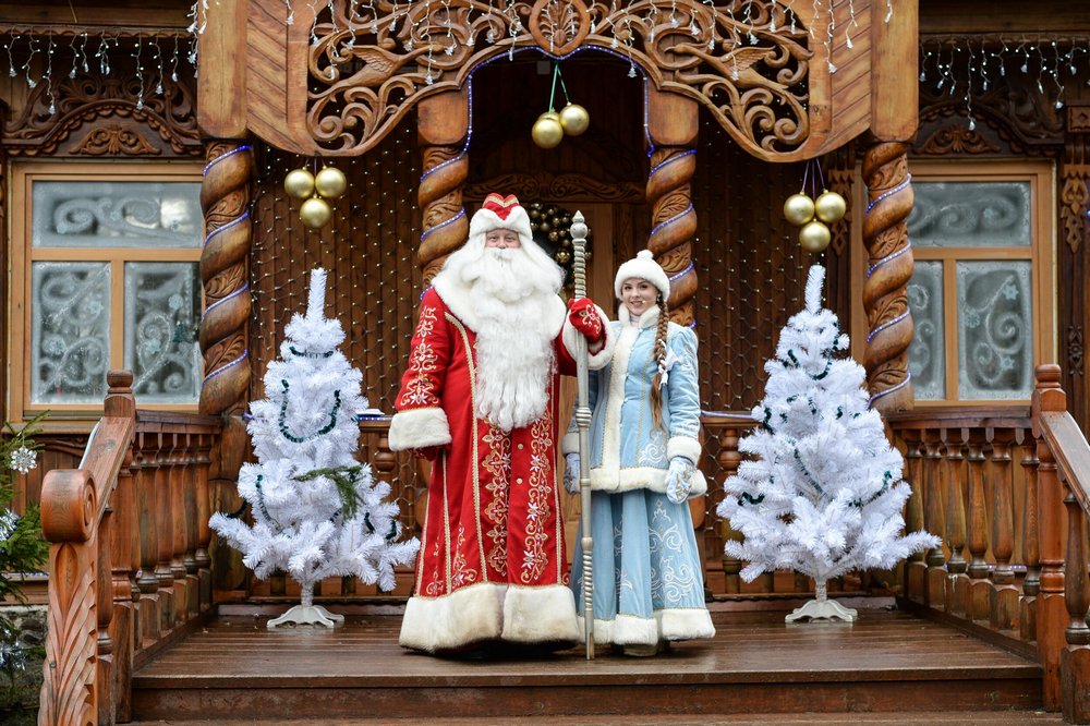 Дед Мороз и Снегурочка на фоне деревянного дома, искусственные ели